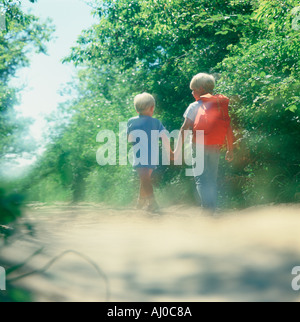 Two children walk hand in hand down a dusty path by a woods One child is wearing an orange backpack Stock Photo