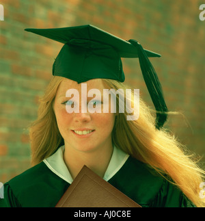 Outdoor portrait of a smiling red haired freckled young woman wearing a cap and gown for graduation Stock Photo