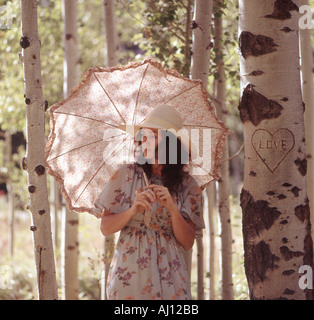 Attractive young woman carrying a parasol strolls through forest setting Carved on a nearby tree is a heart with the word love Stock Photo