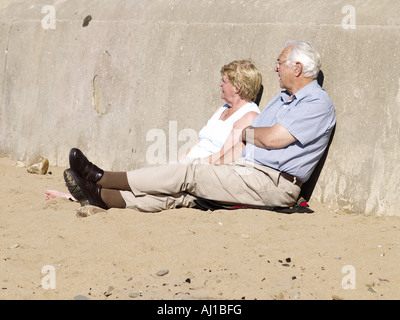 middle aged couple leaning against a sea wall, on a sandy beach Stock Photo