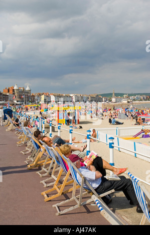 A row of holiday makers on deckchairs on the promenade in front of the crowded sandy beach at Weymouth. Stock Photo