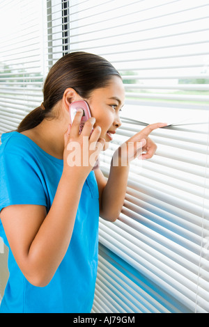Side view of Asian preteen girl looking through blinds out window while talking on cell phone