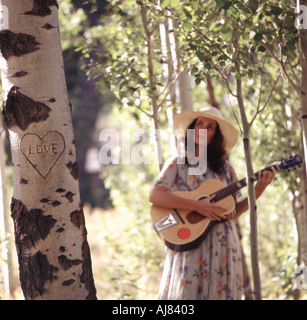 Attractive young woman carrying a guitar strolls through forest setting carved on nearby tree is heart with word love Stock Photo