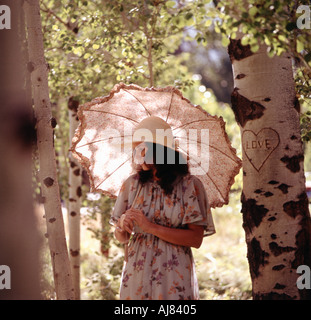 LAttractive young woman carrying a parasolo strolls through forest setting Carved on a nearby tree is a heart with word love Stock Photo