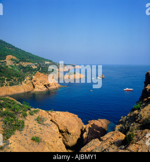Looking across rugged rocks into blue bay with small motor launch at anchor Cap Roux Corniche de l`Esterel in South of France