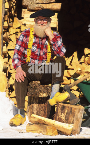 bearded dairyman sitting on log in front of wood pile, wearing beret and wooden shoes Stock Photo