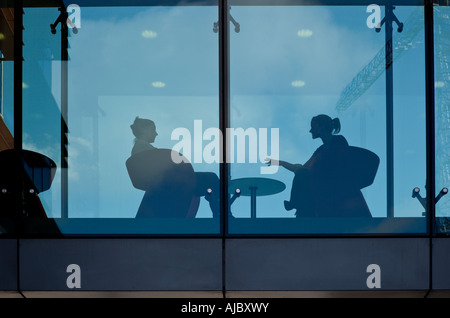 two women talking during a break in the working day are seen seated and silhouetted behind glass Stock Photo