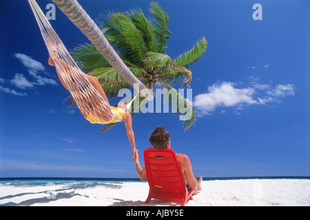 Couple chilling out in hammock and chair under palm tree and sunny skies on white sandy beach on Fihalhohi Island in Maldives Stock Photo