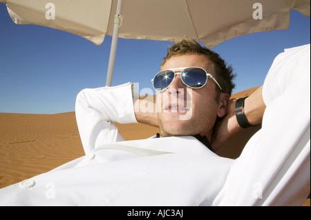 Man in a White Suit and Sunglasses Relaxing Under an Umbrella in the Desert Stock Photo