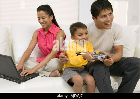 Father playing video game with son by mother using laptop on couch Stock Photo