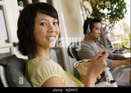 Woman having breakfast on porch, man using laptop in background Stock Photo