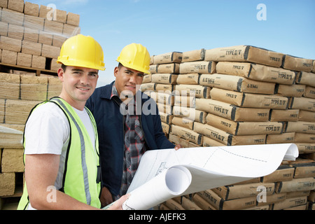 Construction Workers standing near supplies, wearing hard hats, looking at plan on Site Stock Photo