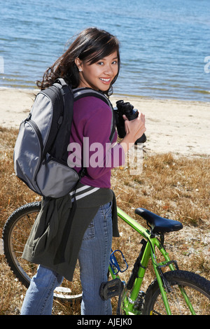 Young woman standing with mountain bike using binoculars by lake Stock Photo
