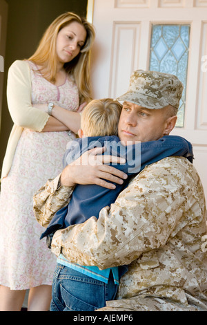 Military father embracing son (5-6) outside home, wife watching Stock Photo