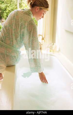 Woman in bathrobe bending down over bathtub filled with bubbles, testing water, back view Stock Photo