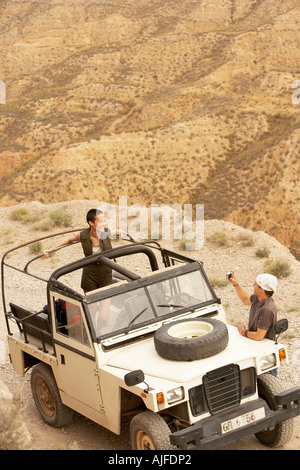 Man photographing woman standing in four wheel drive car on edge of cliff, elevated view Stock Photo