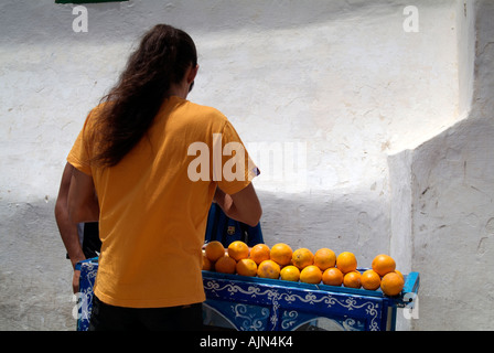 Man with his back to the camera is selling freshly squeezed orange juice from his small blue cart which is piled with oranges. Stock Photo