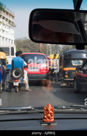 A small Ganesh on a car dashboard. Road traffic is seen through the windscreen. A reflection of a chauffeur in the rear view mirror.  Mumbai, India Stock Photo