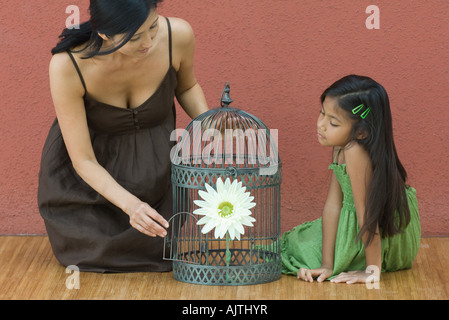 Mother and daughter on floor on either side of birdcage containing flower, woman opening door of birdcage Stock Photo