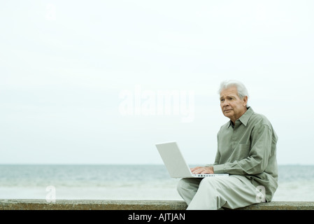 Senior man sitting near water's edge, using laptop Stock Photo