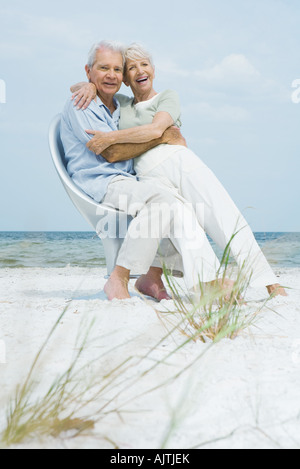 Senior couple sitting in chair together on beach, embracing, woman sitting on man's lap, both smiling at camera Stock Photo