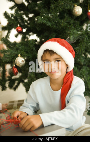Boy opening present in front of Christmas tree, wearing Santa hat, smiling at camera Stock Photo