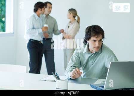 Businessman working on laptop, colleagues standing in background Stock Photo