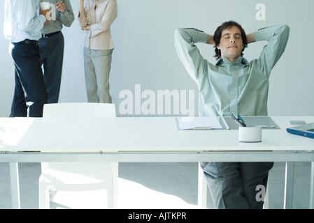Businessman sitting at desk with hands behind head and eyes closed, colleagues standing in background Stock Photo