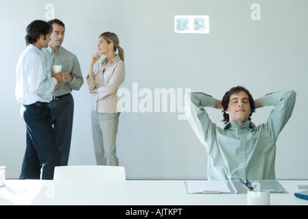 Businessman sitting at desk with hands behind head and eyes closed, colleagues standing in background Stock Photo