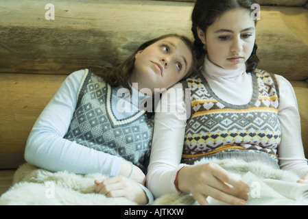 Two teen girls in winter clothes sitting under warm blanket together, one reading while other looks bored Stock Photo