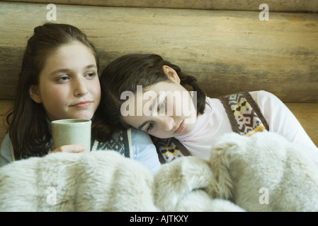 Two teen girls in winter clothes sitting under warm blanket together, one holding hot beverage Stock Photo