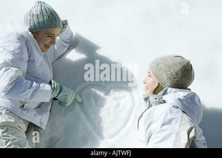 Teenage girls lying on snow, laughing, heart with initials drawn on surface of snow Stock Photo