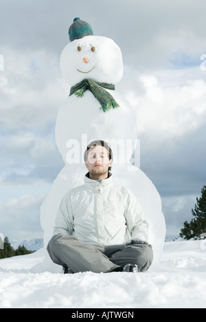 Young man sitting with legs crossed and eyes closed in front of snowman Stock Photo