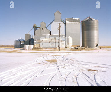 Tire tracks in snow lead up to a steel grain elevator in the snow-covered prairies of South Dakota, against a cloudless blue sky Stock Photo