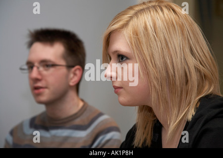 blonde haired young woman sitting beside dark haired young man wearing glasses listening to someone Stock Photo