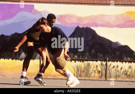 Two young men play basketball on a playground whose wall is a painted mural Stock Photo