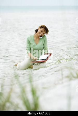Woman sitting on sand, using cell phone and writing in agenda Stock Photo