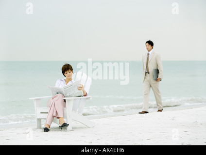 Woman sitting reading newspaper on beach as businessman carrying laptop approaches Stock Photo