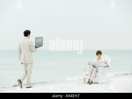 Woman sitting reading newspaper on beach as businessman carrying open laptop approaches Stock Photo