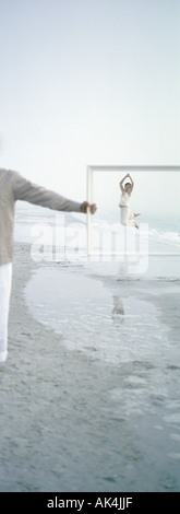 On beach, woman jumping in distance, seen through frame held by man Stock Photo