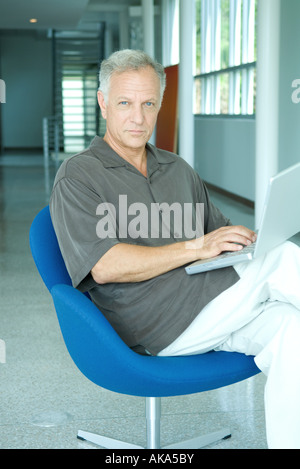 Mature man sitting in chair, using laptop computer, looking at camera, portrait Stock Photo
