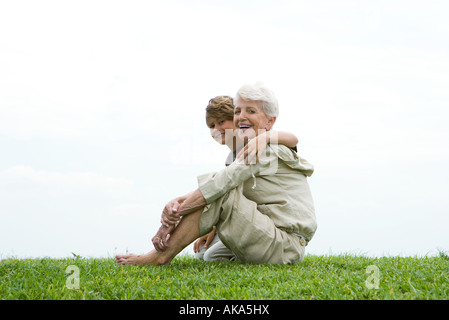 Senior woman and grandson sitting on grass, smiling at camera, full length