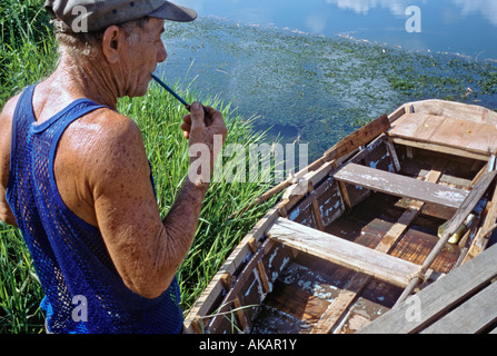 Old man smoking pipe looking at flat bottom boat in the Rio Duaba near Baracoa Cuba Stock Photo