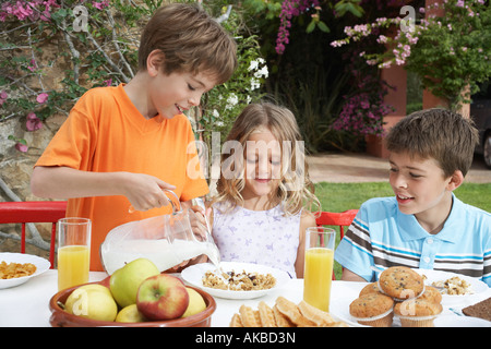 Two boys and girl (6-11) at table, boy pouring milk on cereal, garden Stock Photo