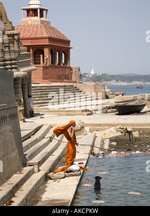 A man in colouful robes on the banks of the Ganges. He is preparing to join his dog cooling down in the river. Varanasi, India. Stock Photo