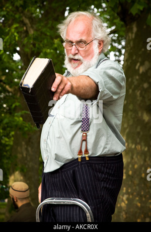 A white-bearded Christian preacher at Speaker's Corner pointing with a bible in hand. A Muslim man is seen behind. Stock Photo