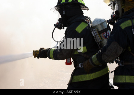 Two firefighters holding a hoseline putting water onto a fire at an emergency scene with smoke and stream all around them Stock Photo