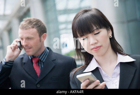 Close-up of a businesswoman using a personal data assistant with a businessman talking on a mobile phone behind her Stock Photo