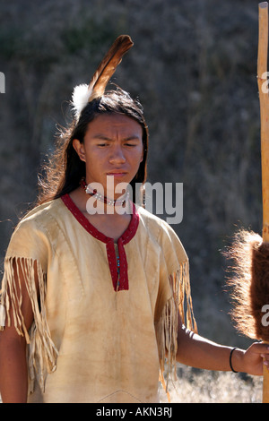 A Native American Indian boy wearing a feather standing next to a horse holding a coup stick with buffalo hide on it Stock Photo