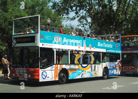 Tourist passengers seated top deck of two open top double decker tour buses outside 'Spanish Village' retail shopping & cafe complex Barcelona Spain Stock Photo